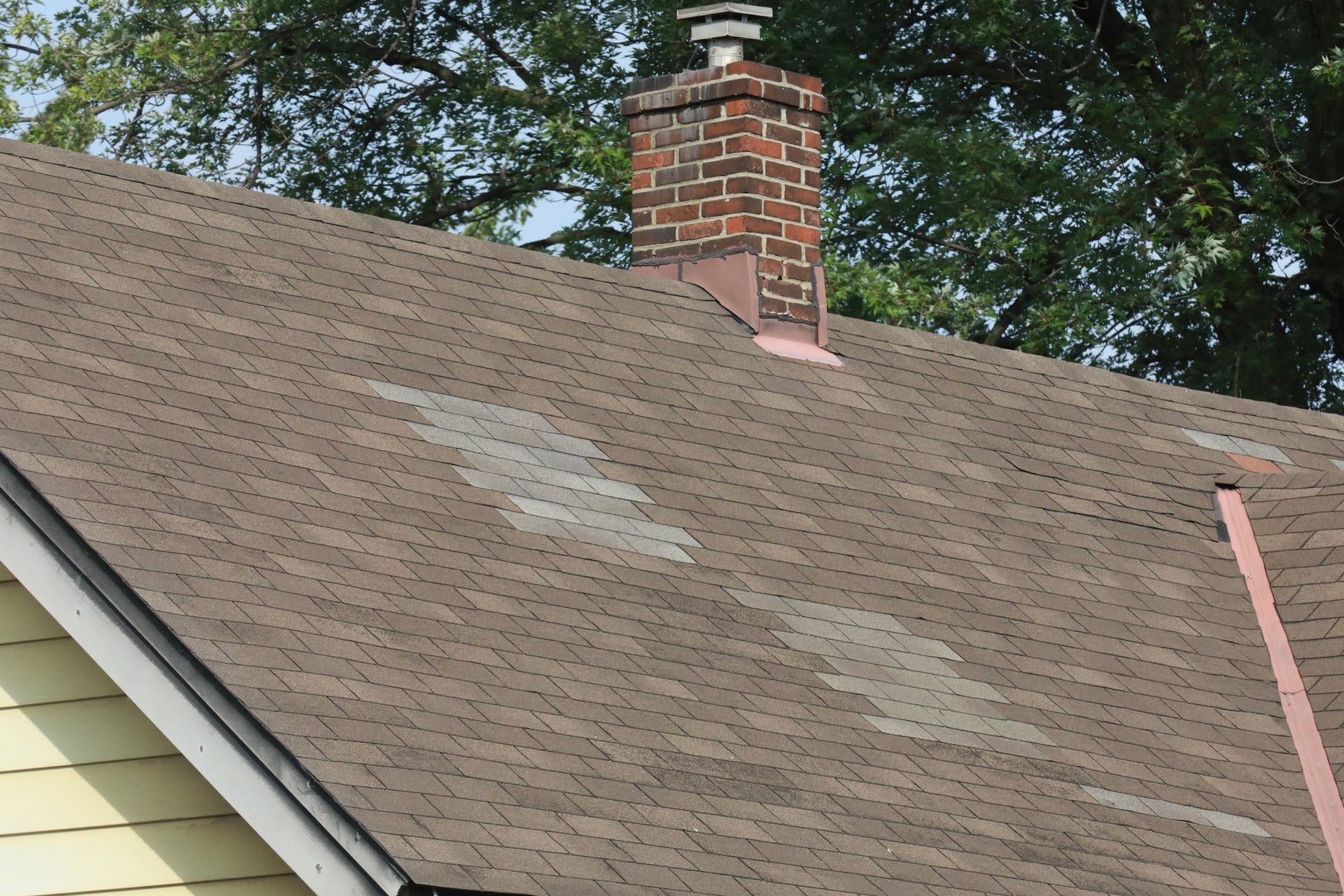 House roof with scattered light-colored patches and a brick chimney under trees