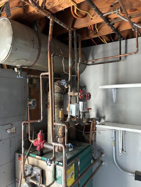 Boiler room with large metal tank, pipes, pumps, and exposed ceiling beams in an industrial basement.