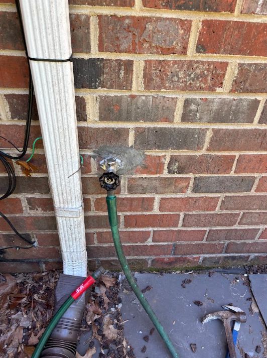 Outdoor brick wall with a water spigot and green garden hose attached beside a downspout.