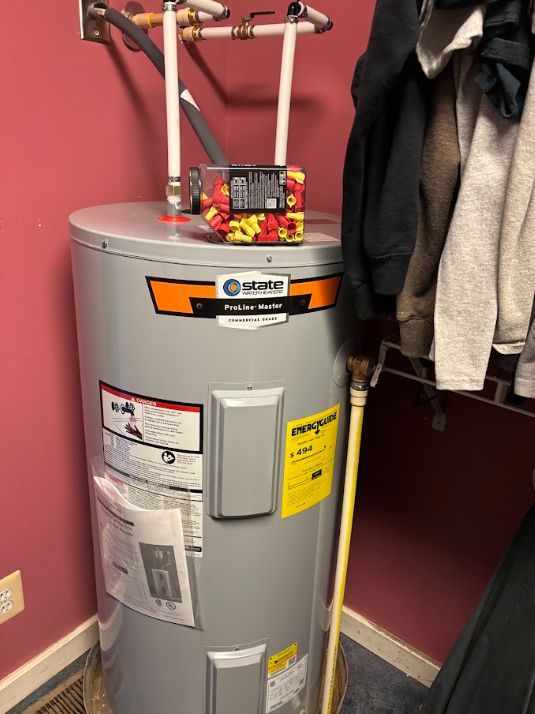 Water heater in a closet with clothes hanging nearby, red wall and gray tank.