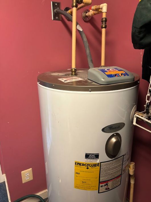 White water heater against a pink wall with exposed plumbing and nearby outlet, in a utility closet
