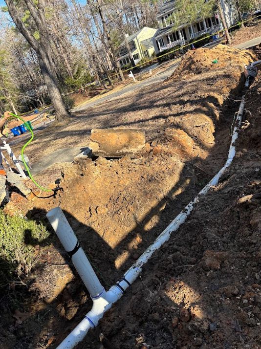 Backyard trench with white PVC pipe laid along a dirt path beside a house and trees.