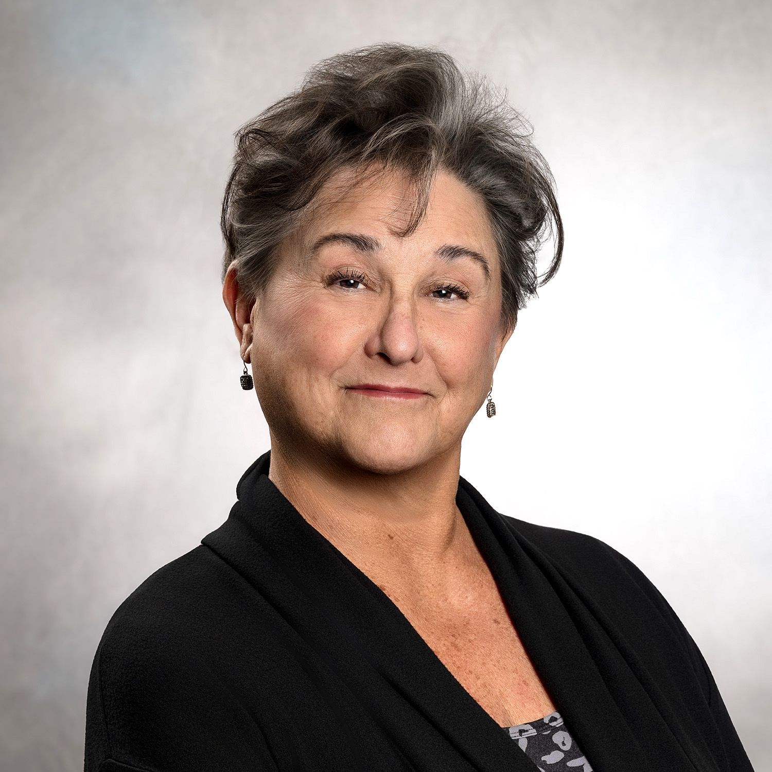 Woman with short gray hair, wearing a black cardigan, smiles at the camera. Neutral background.