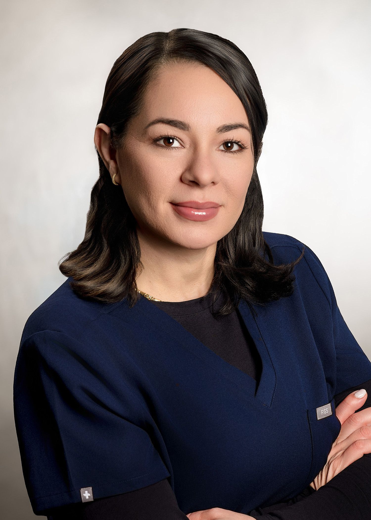 A professional portrait of a person with dark hair wearing dark blue scrubs, arms crossed, smiling slightly.