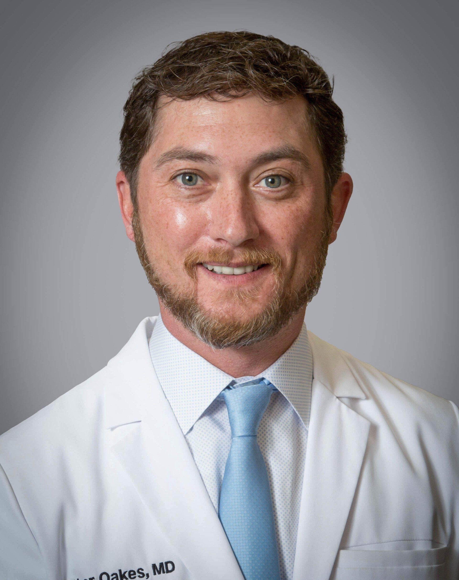 A man with a beard is wearing a white coat and blue tie.