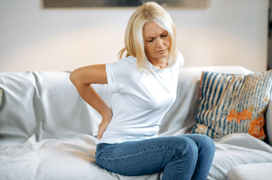 A woman is sitting on a couch holding her back in pain.