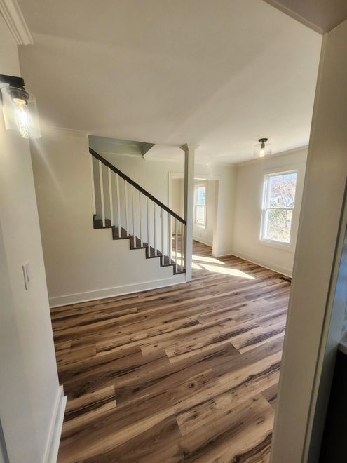 Empty living room with wood floors, staircase, and bright windows.