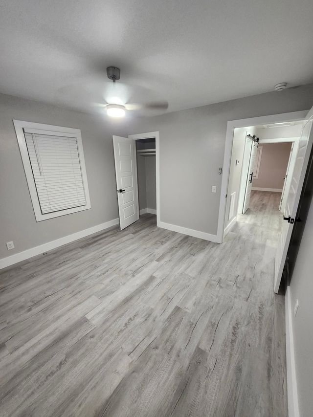 Empty gray bedroom with light wood floors, ceiling fan, open closet, and white blinds.