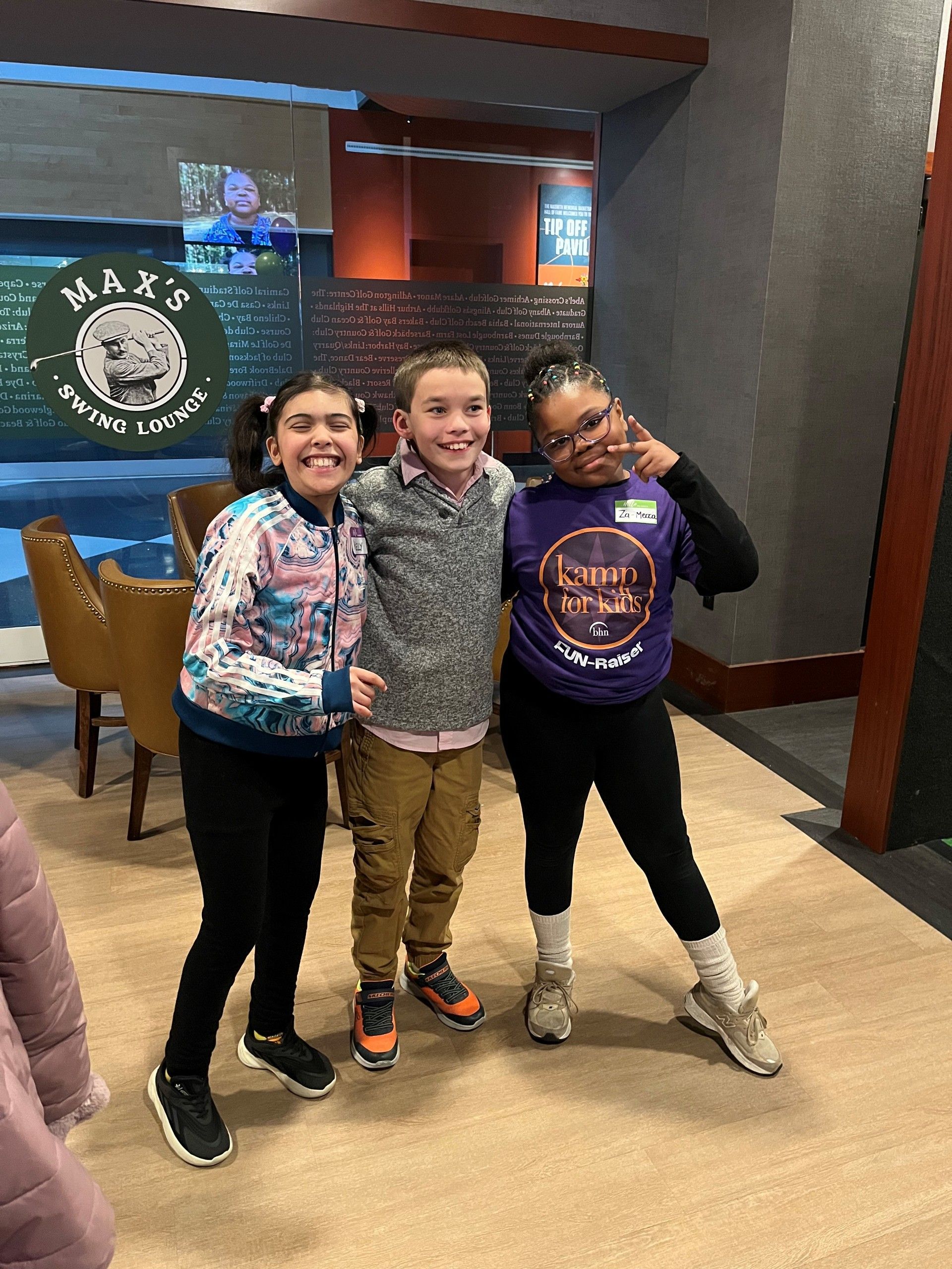 Three smiling kids pose together indoors, standing arm-in-arm in front of a restaurant sign.