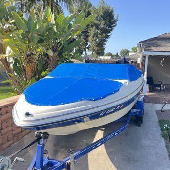 A white and blue boat on a trailer, covered with a blue tarp, parked in front of a house on a sunny day. A white and blue boat on a trailer, covered with a blue tarp, parked in front of a house on a sunny day.