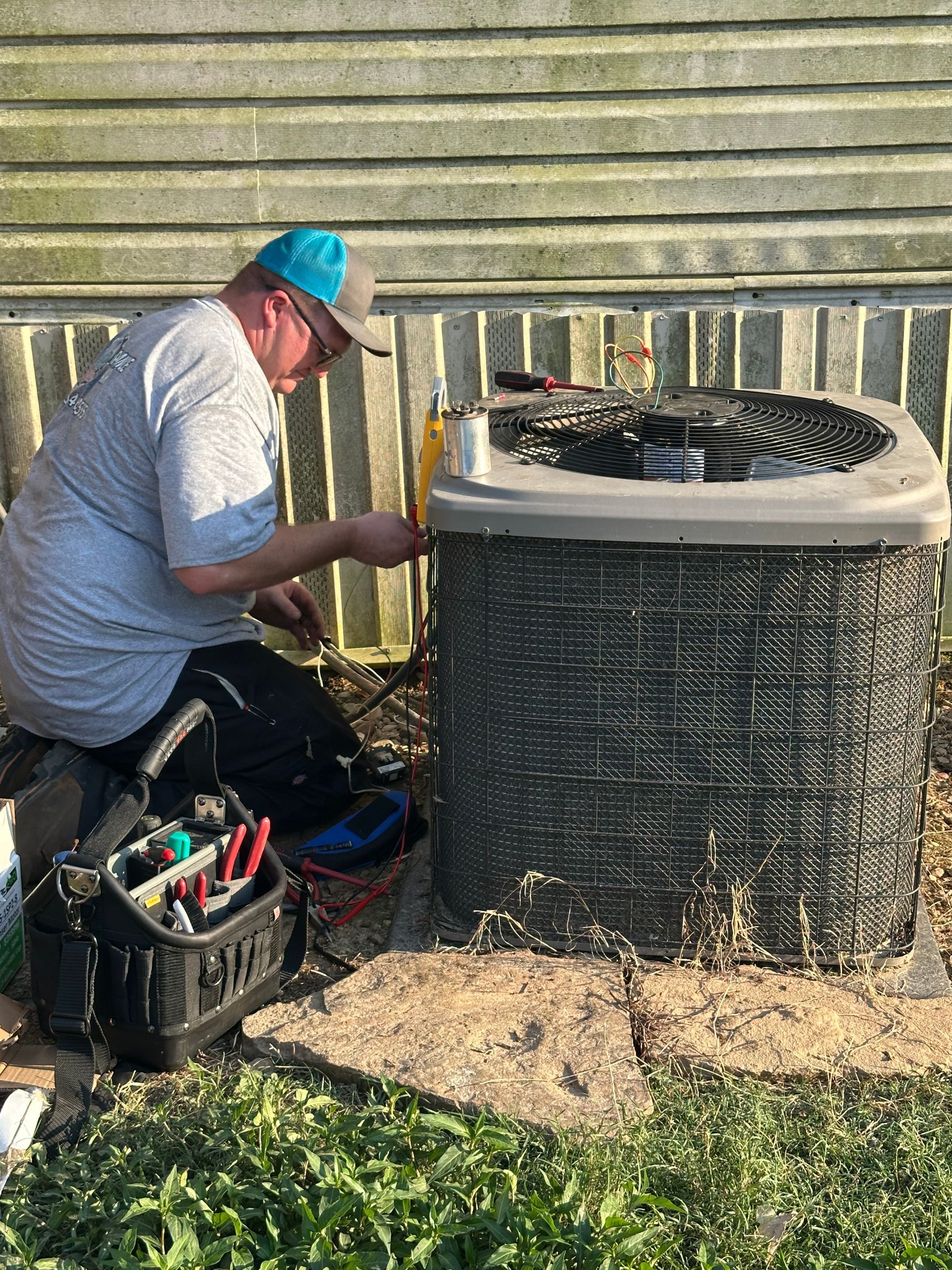 Technician kneels beside an outdoor HVAC unit, using tools to repair it against a house wall.