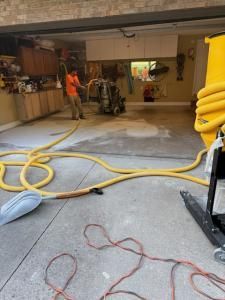 A person in an orange shirt uses a water extraction machine in a garage, with yellow hoses and equipment.