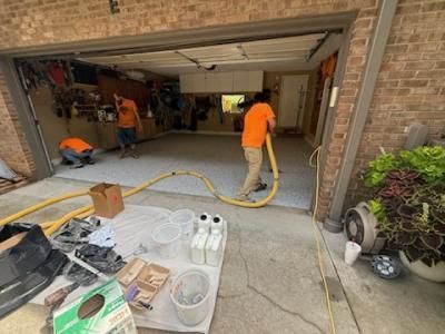 Workers applying flooring material in a garage. Men in orange shirts work with hoses and equipment; supplies sit nearby.