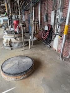 Industrial factory floor with machinery and a large, round manhole cover. Brick wall in background.