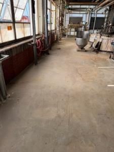 A long, empty industrial hallway with concrete floor, windows, and metal equipment.