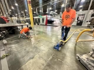 Two workers repairing a concrete floor in a large industrial setting. One uses a floor grinder, the other works on the floor with hand tools.
