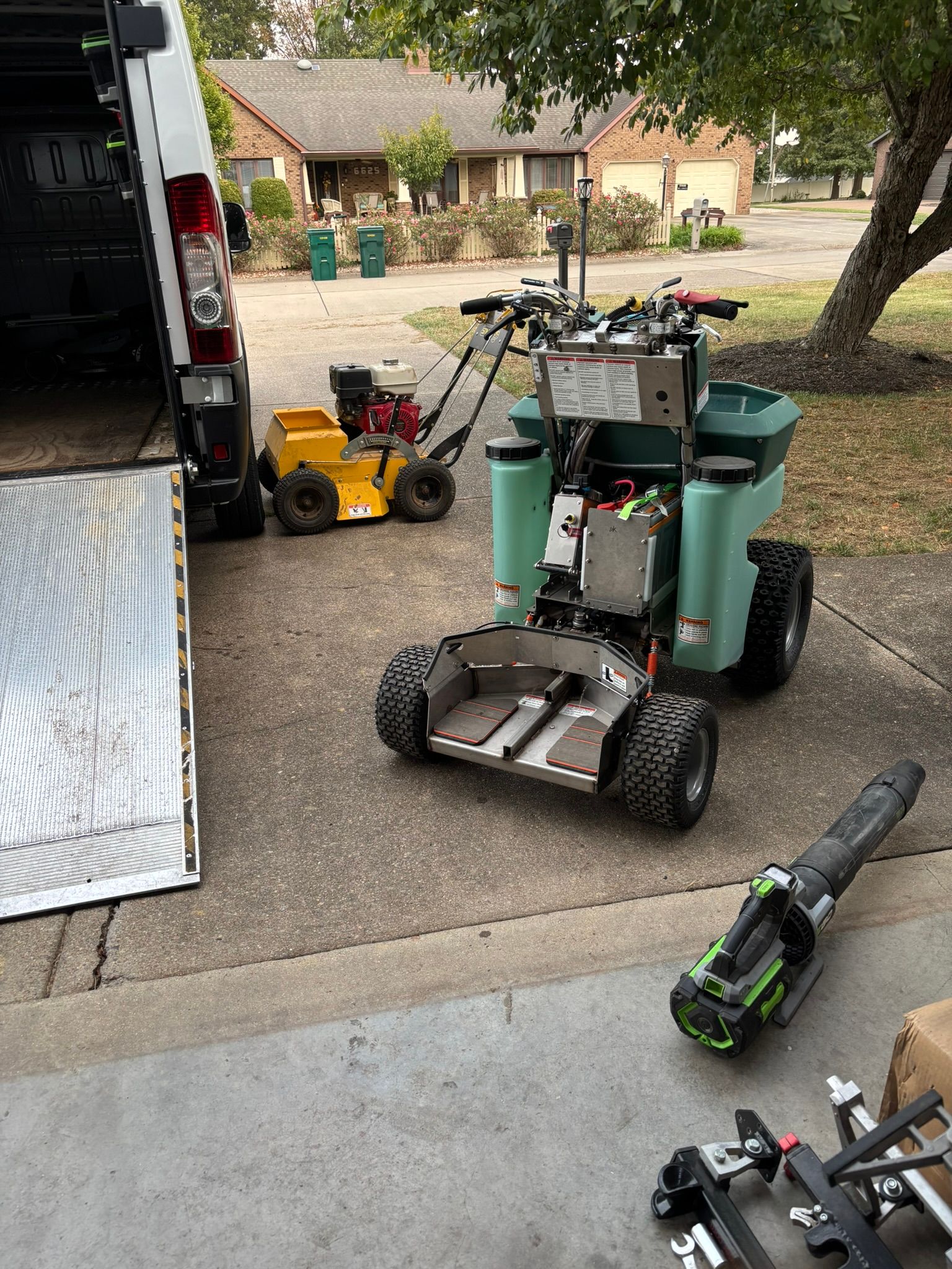 Lawn mowers and outdoor tools stored on a driveway beside a truck ramp and yard.