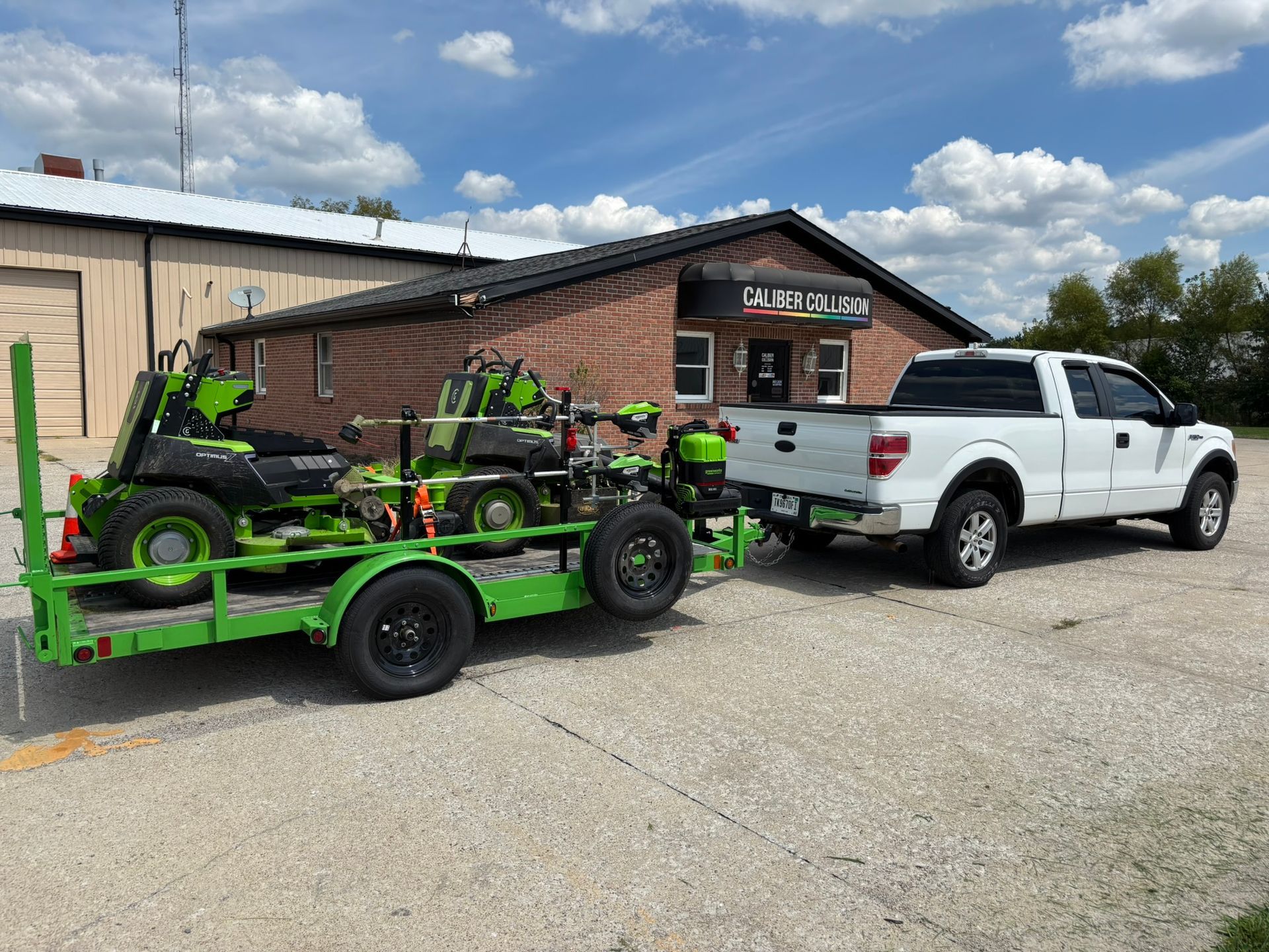 White pickup truck towing a green lawn mower trailer outside a brick building under a cloudy sky