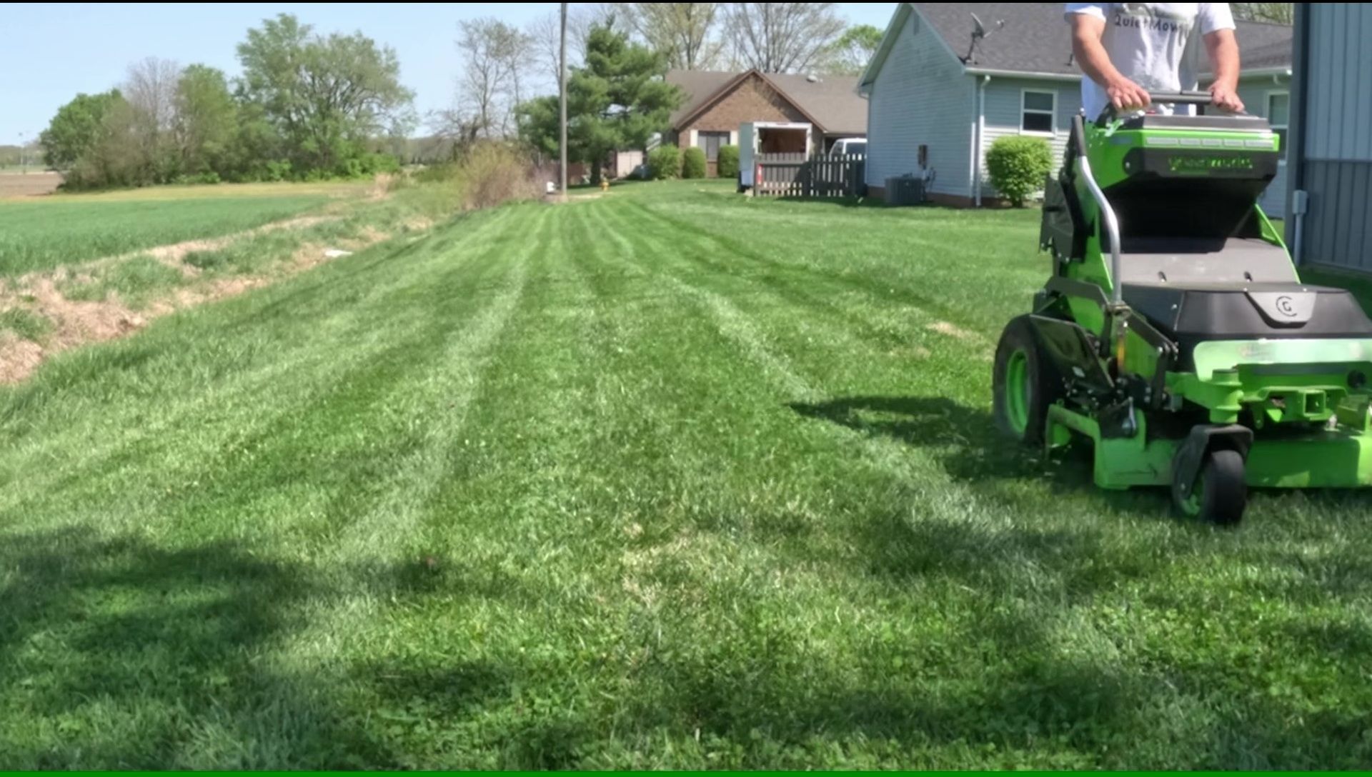 Person mowing a lawn with a green riding mower; cut grass and trees in the background on a sunny day.