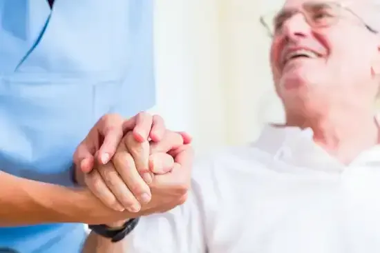 Une soignante tient les mains d'une personne âgée ; ils sourient dans une pièce baignée de lumière.