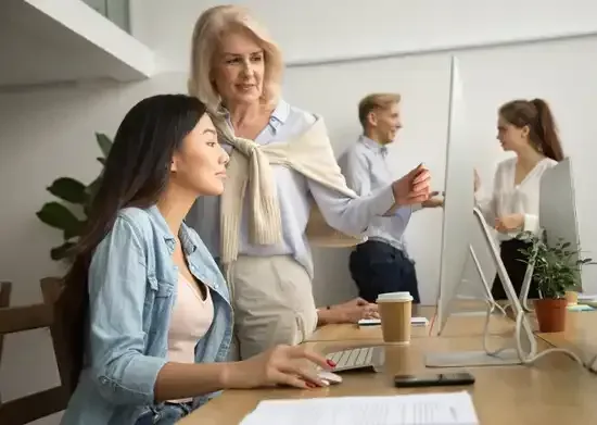 A woman is standing next to a woman sitting at a desk in front of a computer.