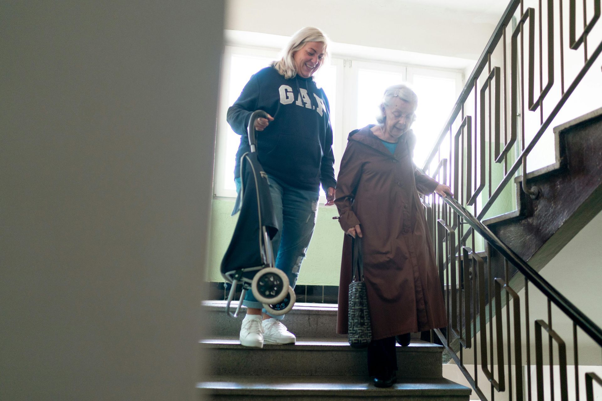 Two people and a small dog stand on an indoor staircase near a bright window.