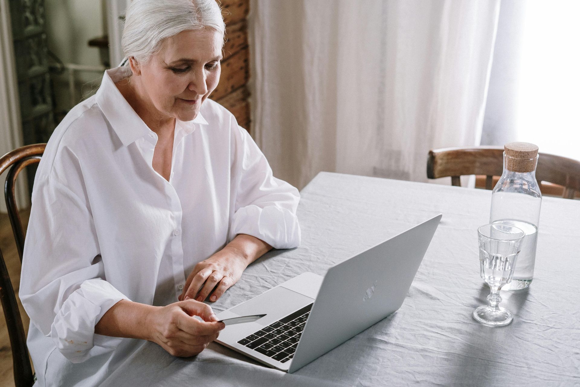 Person using a laptop at a dining table beside a glass bottle of water, wearing a white shirt and head wrap.