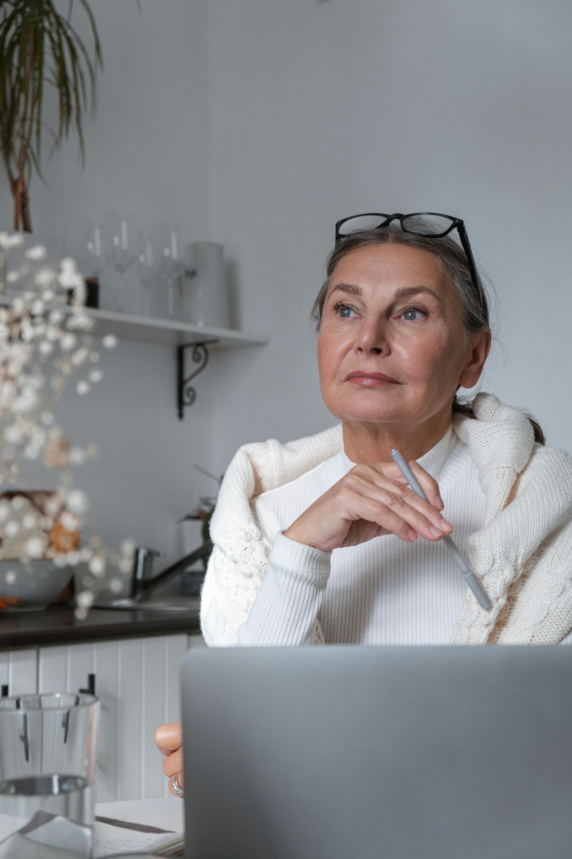 Woman sitting at a laptop in a bright kitchen, resting her chin on her hand and looking thoughtfully ahead