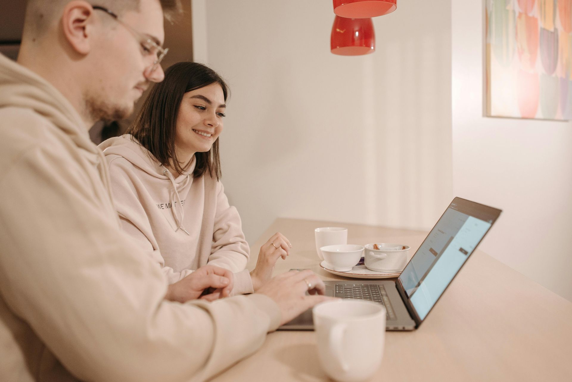Two people sitting at a table using a laptop, with coffee cups in a bright room.