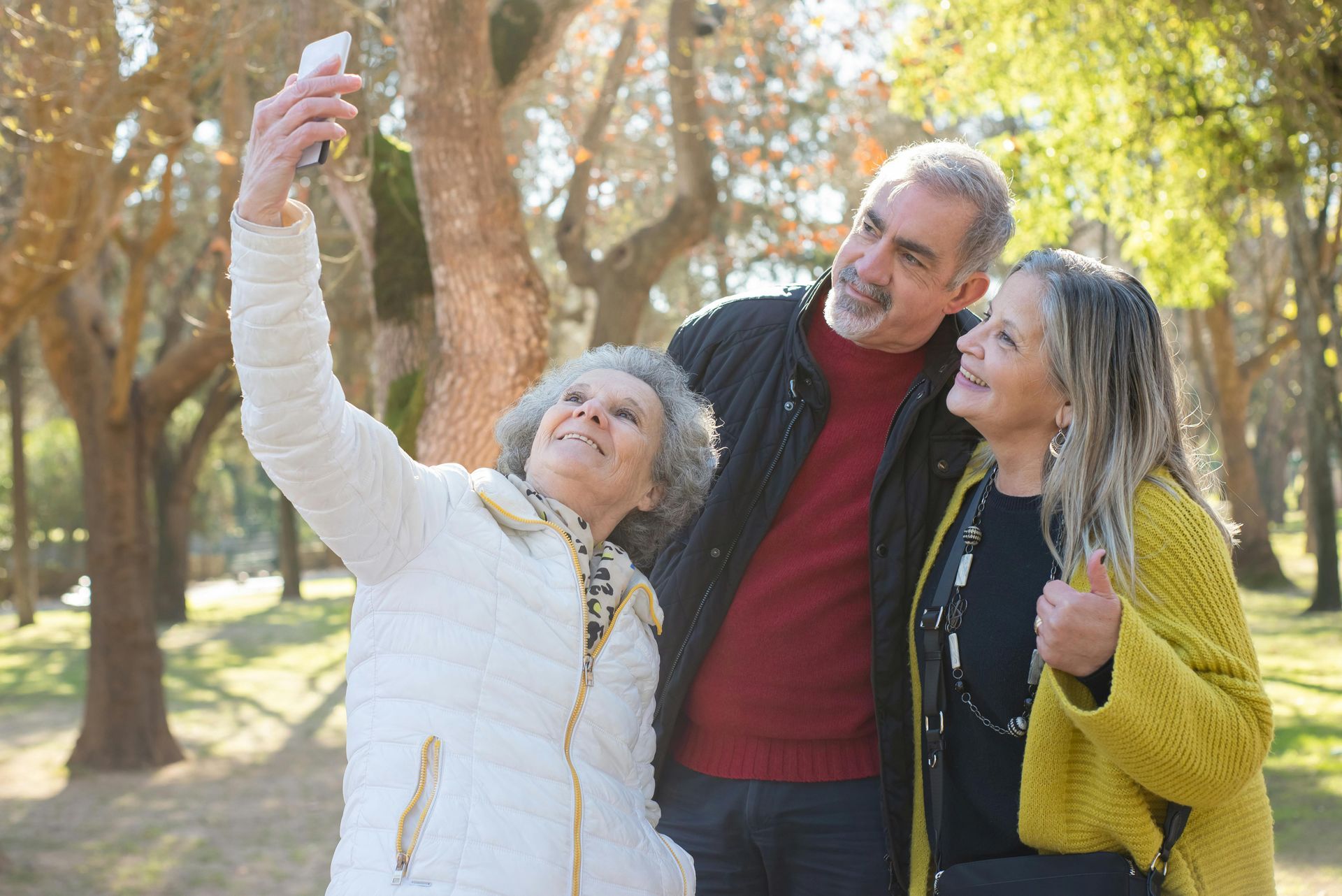 Three people smiling outdoors, taking a selfie among trees in a sunny park