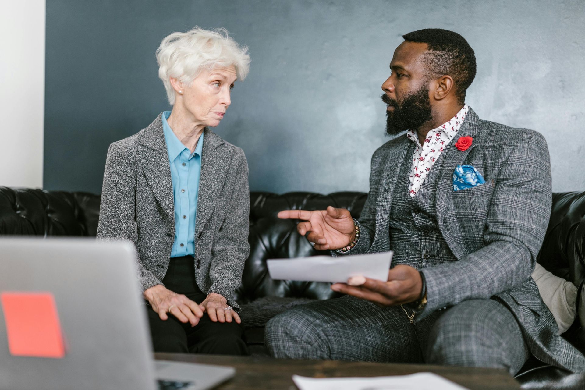 Two coworkers in suits talking on a couch, one holding a tablet in a modern office.