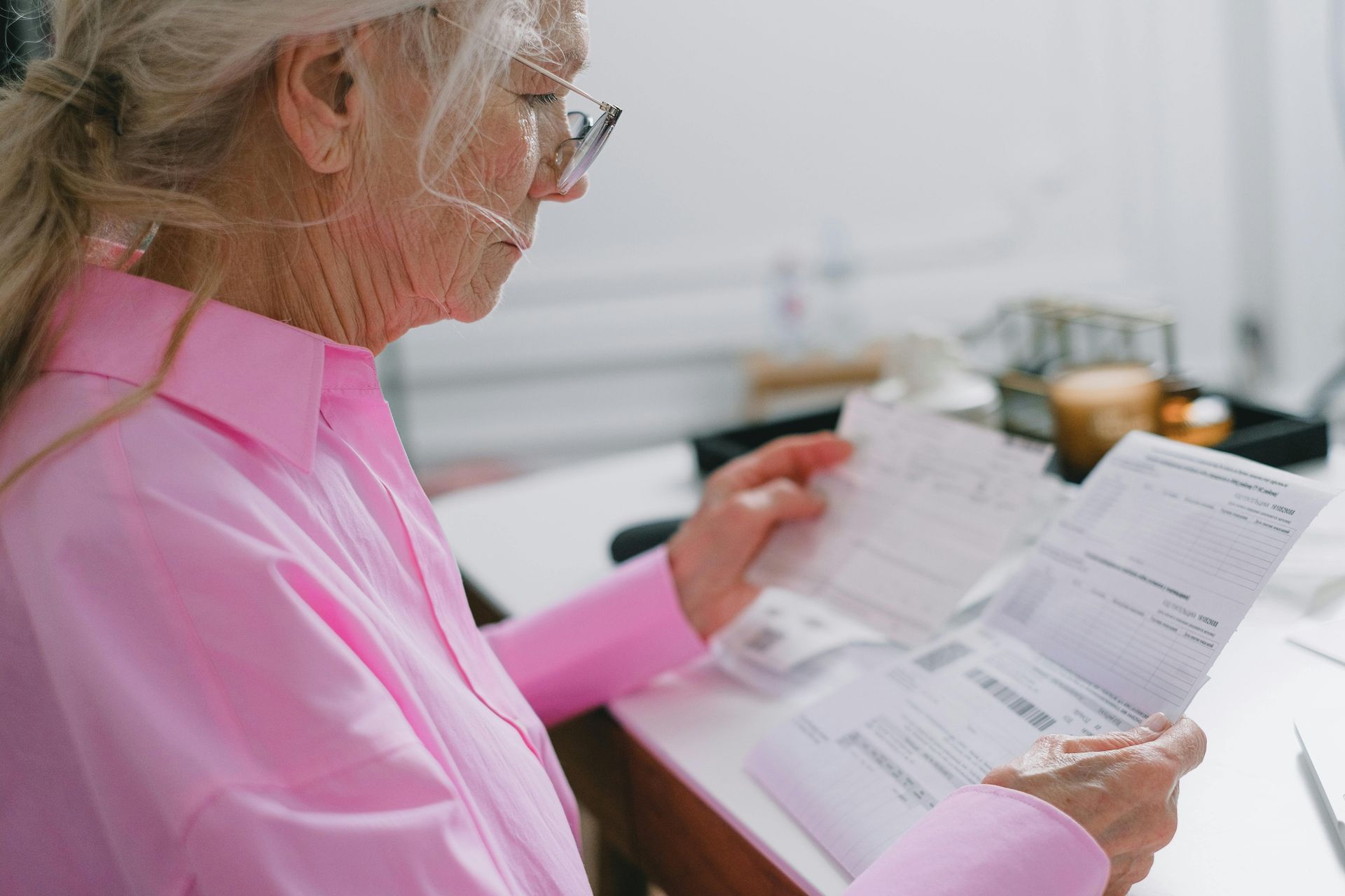 Person in a pink shirt reading paperwork at a desk with a laptop and office supplies