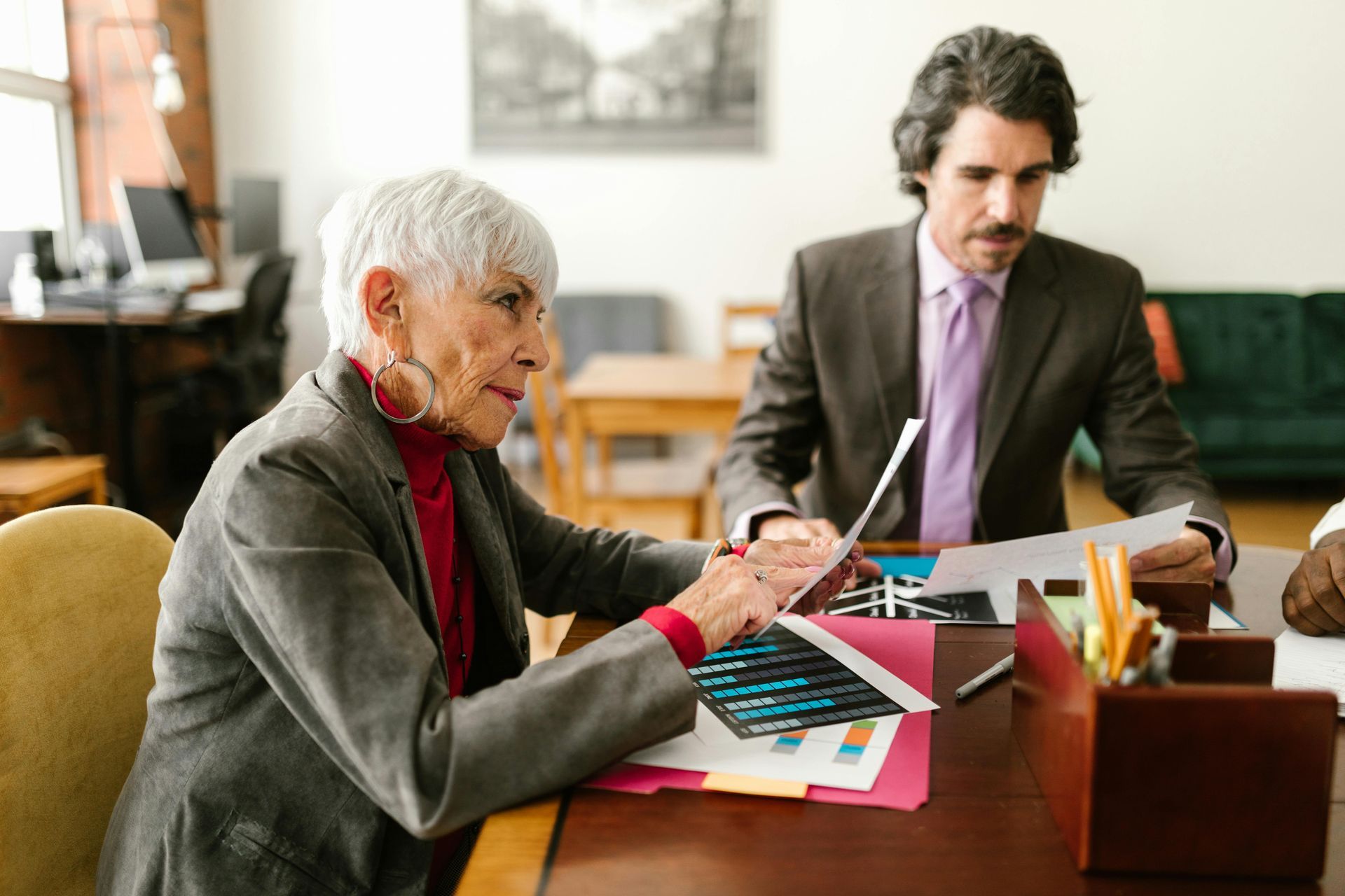 Two people reviewing documents at a desk in a bright office, with papers, a laptop, and stationery nearby