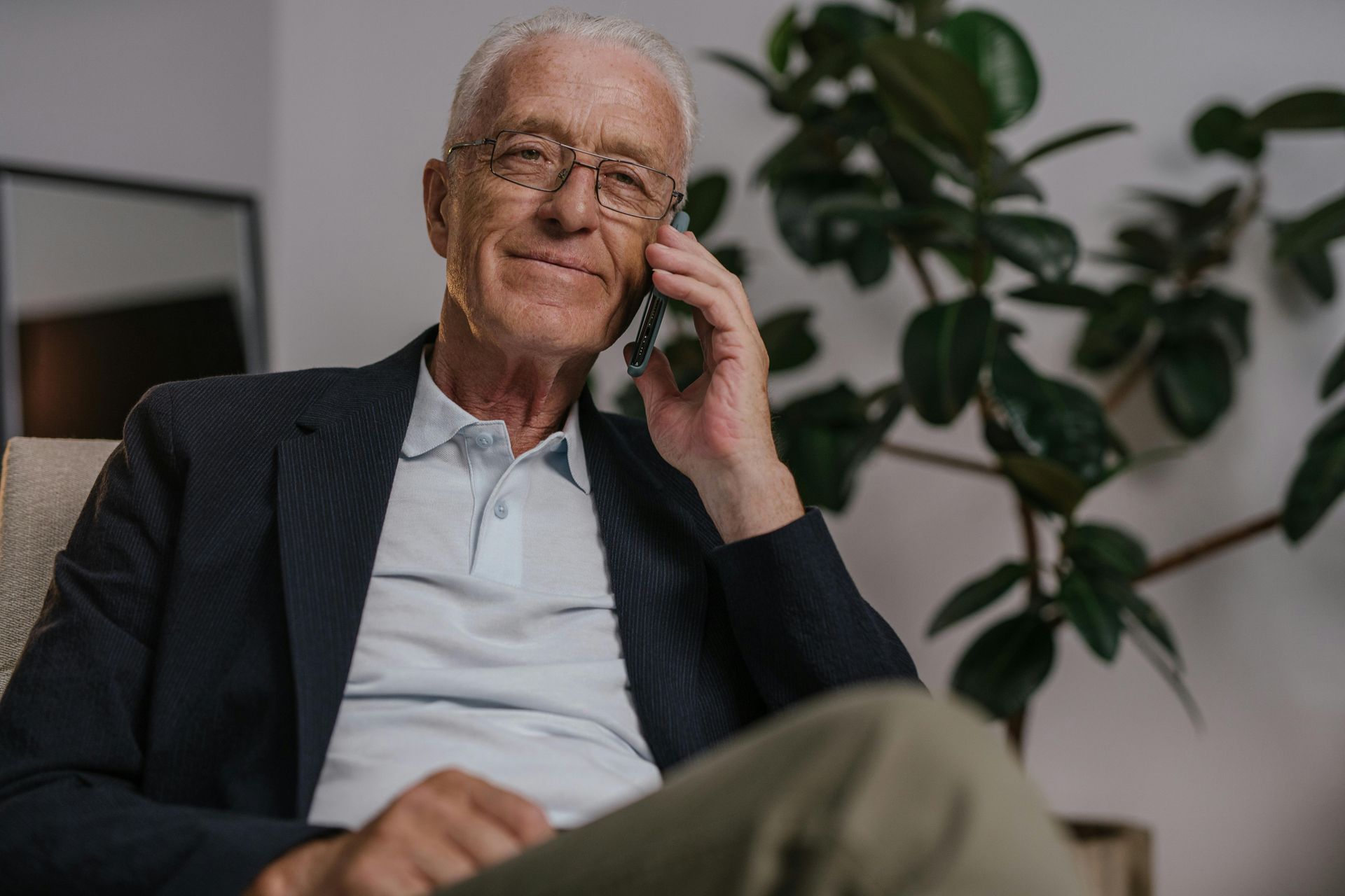 Older man in a dark blazer sitting indoors, smiling while talking on a phone with a plant in the background