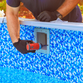 Gloved hands using a drill to install a pool wall fitting on a blue-lined pool side