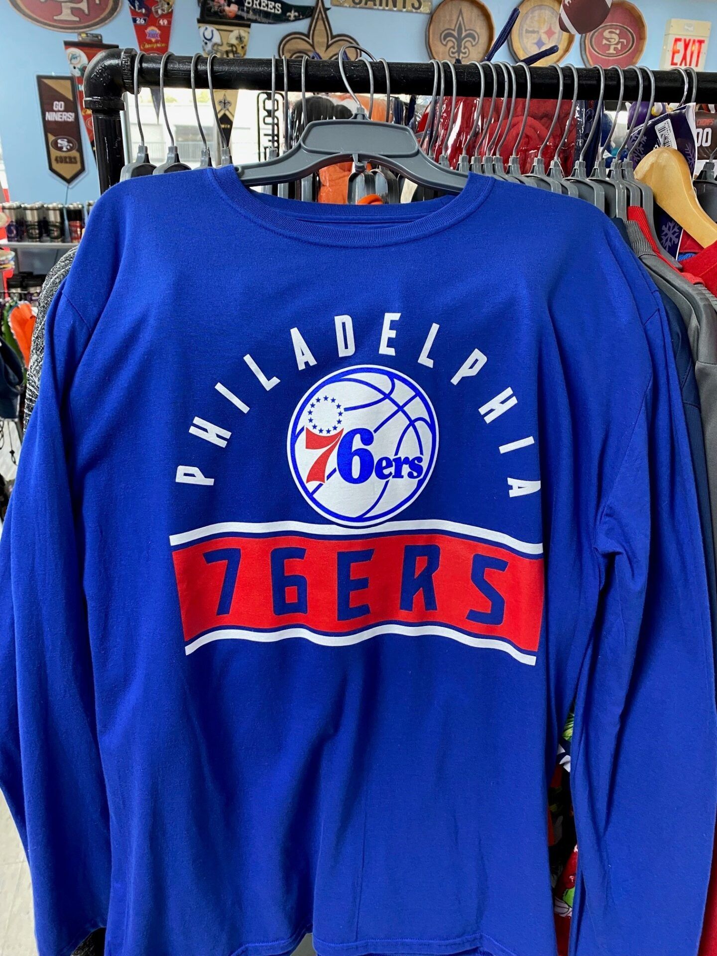 Blue Philadelphia 76ers long-sleeve shirt hanging on a rack in a store.