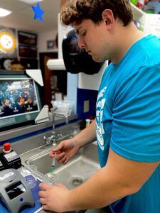 Person washing dishes at a sink beside a laptop in a kitchen-like workspace