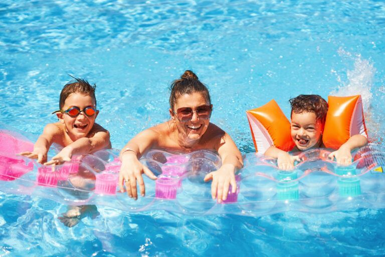 two children and one female adult smiling and floating in a bright blue swimming pool with pink and orange floaties