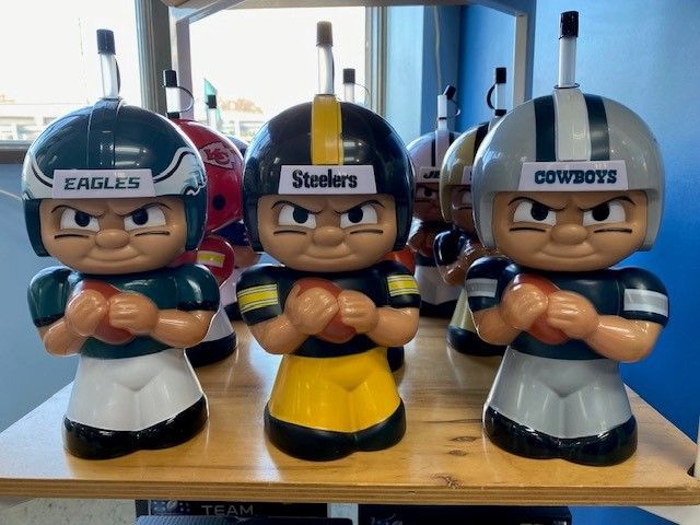 Three football helmet figurines on a table with arms crossed, labeled Eagles, Steelers, and Cowboys.