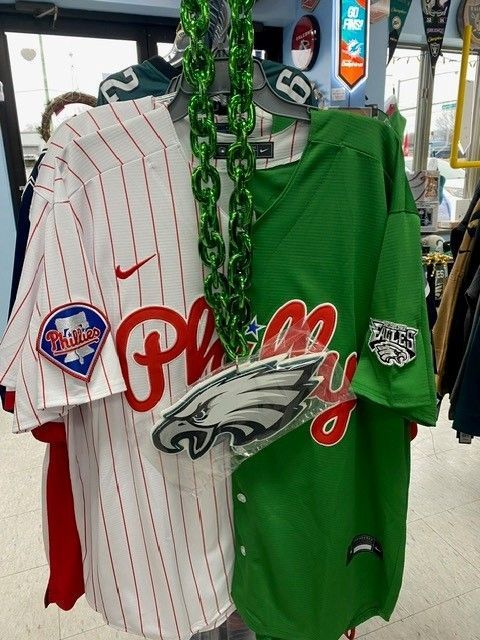 Two Phillies baseball jerseys hanging on a rack, one white pinstripe and one green, with an Eagles logo on the green one