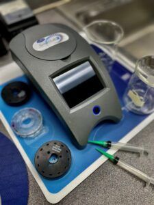 Gray lab device on a blue tray with syringes, tubing, and small sample cups