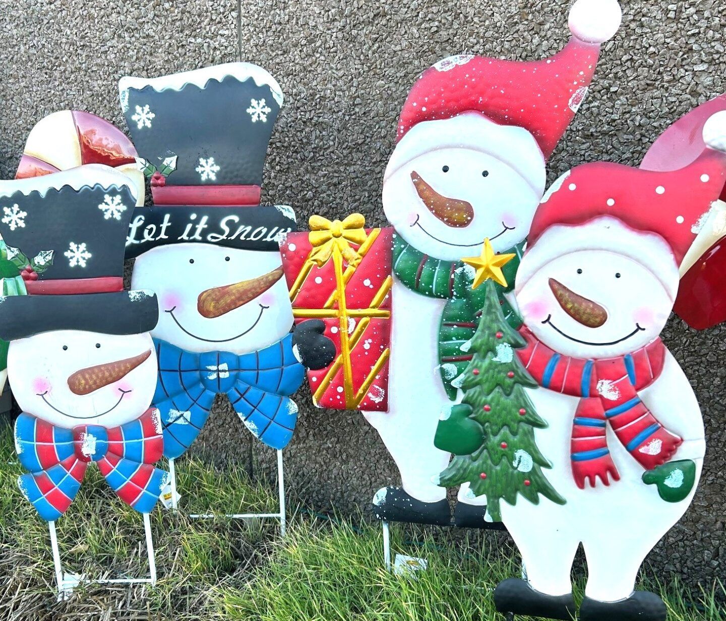 Festive snowman yard decorations with Santa hats, scarves, a tree, and a gift against a stone wall