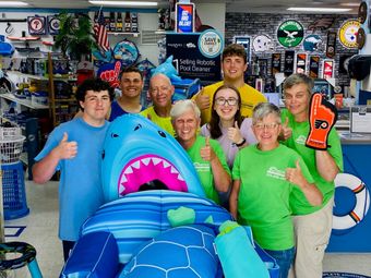 Group posing in a shop behind a large blue inflatable shark display, smiling and holding colorful items.