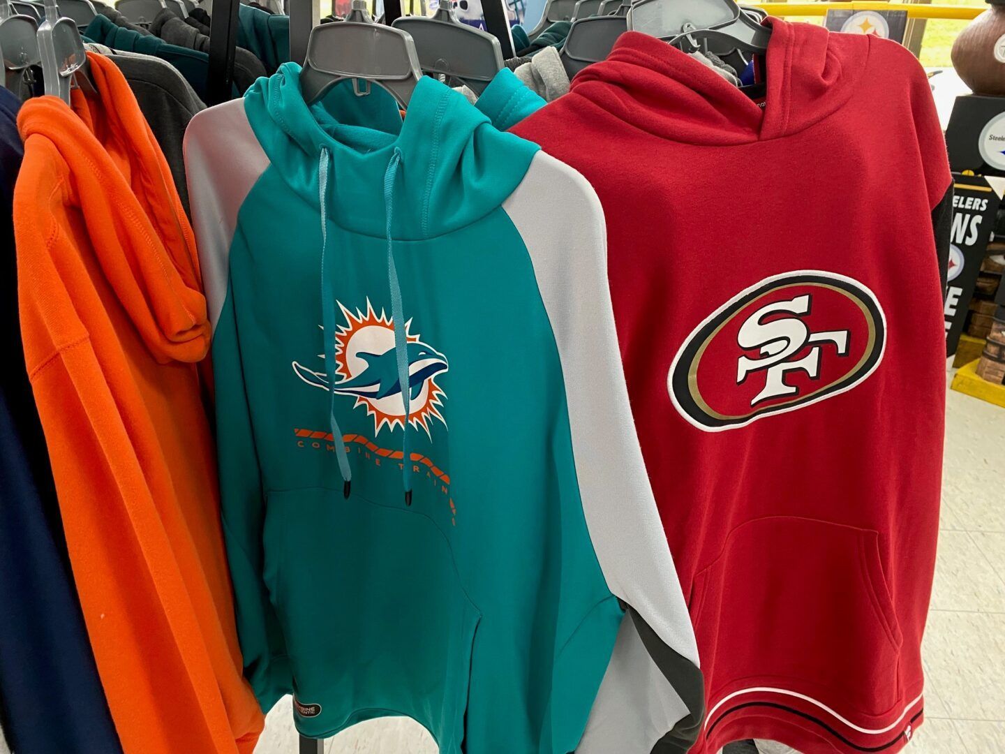 Assorted sports hoodies on a rack, including teal Dolphins, red 49ers, and orange hoodie styles
