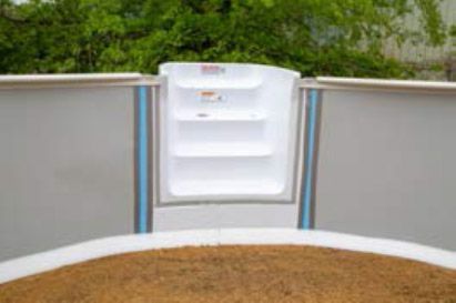 White pool skimmer opening to a yard, with tan ground cover and green trees beyond.