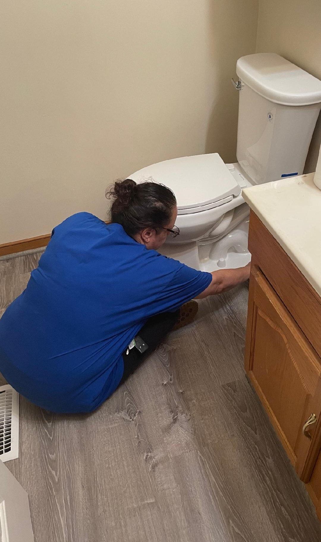 Woman in blue shirt crouches near toilet, possibly repairing it in a bathroom.