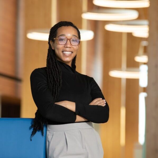 A woman wearing glasses and a black turtleneck is standing with her arms crossed and smiling.
