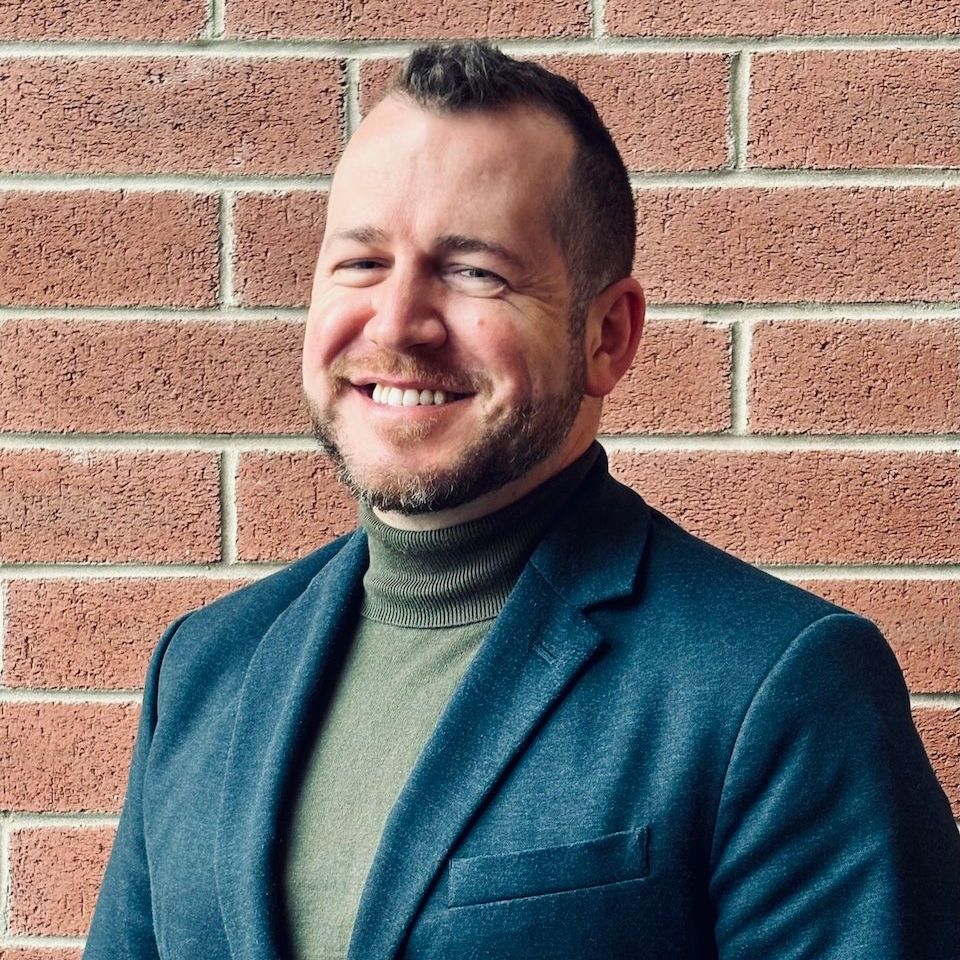 A man in a suit and turtleneck is smiling in front of a brick wall.