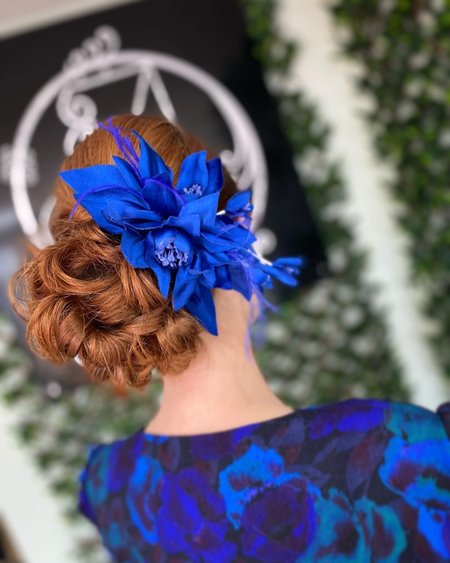 A person with textured auburn hair in an updo, adorned with a large, vibrant royal blue flower accessory.