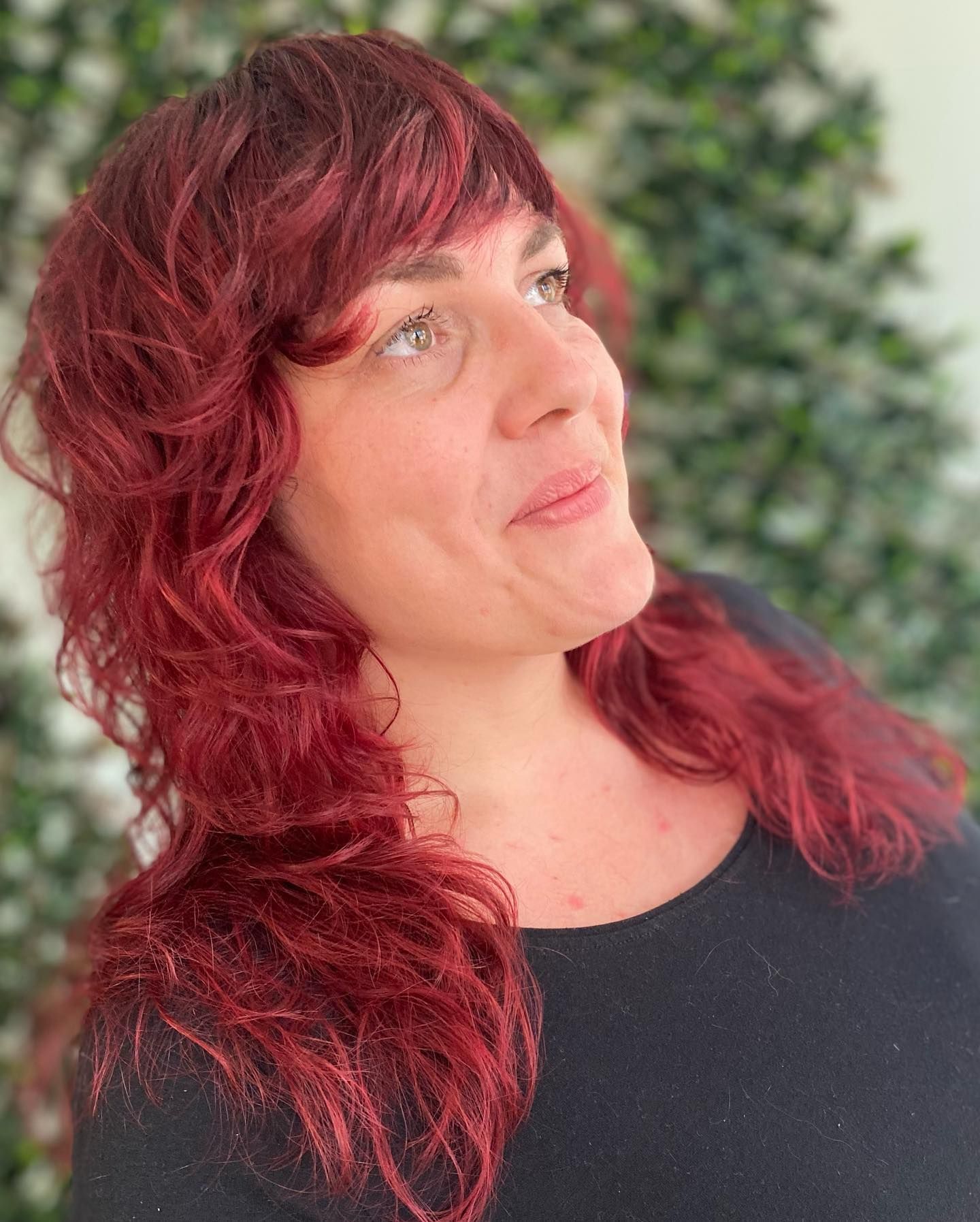 A person with vibrant red layered hair, bangs, and a black top looking upward against a backdrop of green leaves.